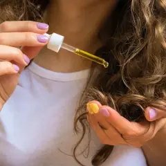 En hand med lila naglar håller en pipett med gyllene rosmarinolja hår. Oljan droppar ner på en lock av brunt, vågigt hår.