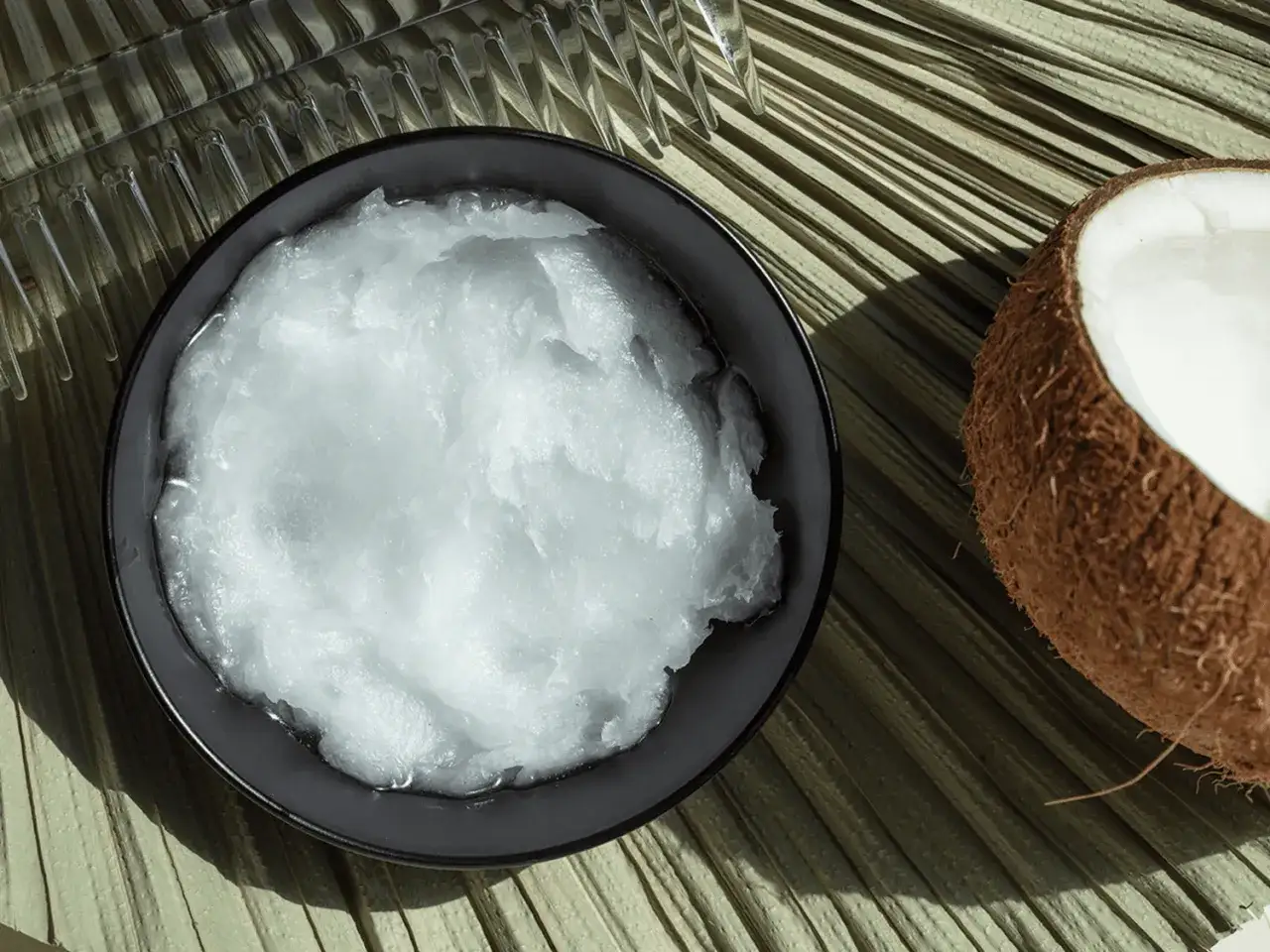 En sk&aring;l med kr&auml;mig kokosolja f&ouml;r huden bredvid ett halvt kokosn&ouml;t p&aring; en palmblad.