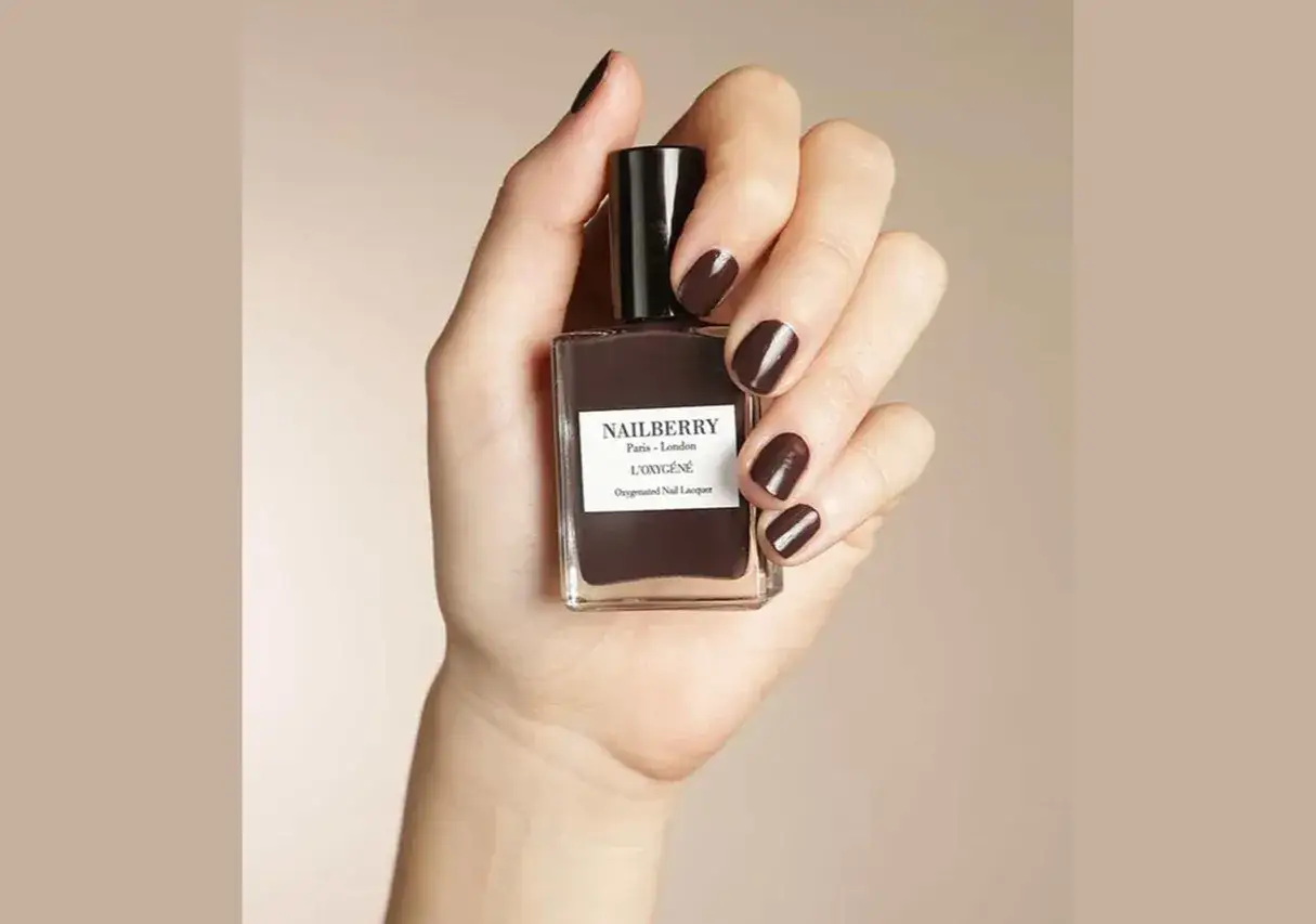 En hand håller en flaska mörkt, chokladbrunt Nailberry nagellack. Naglarna är målade i samma djupa färg.
